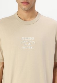 Beige bomull T-skjorte med rund hals, korte ermer og hvit brodert tekst: "GUESS CALIFORNIA L.A. Est. 1981." Glatt tekstur.
