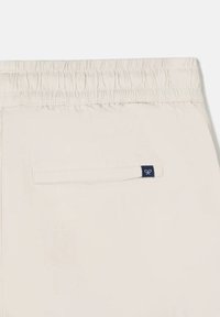 Pantalones beige claros con cintura elástica, cuentan con un bolsillo trasero y una etiqueta azul marino, textura suave y diseño ajustado.