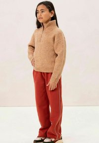 Beiger gerippter Pullover mit hohem Kragen, kombiniert mit roten Jogginghosen, die weiße seitliche Streifen haben. Das Outfit ist locker geschnitten und aus strukturiertem Stoff.