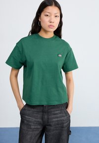 T-shirt verde a maniche corte con colletto rotondo e una piccola patch con logo sul petto sinistro. Indossato con jeans grigi scuri.