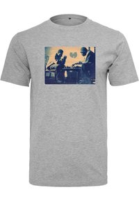 Dos hombres mirándose intensamente mientras juegan al ajedrez, con un logotipo de murciélago estilizado de fondo en una camiseta gris.