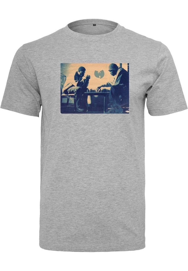 Dos hombres mirándose intensamente mientras juegan al ajedrez, con un logotipo de murciélago estilizado de fondo en una camiseta gris.