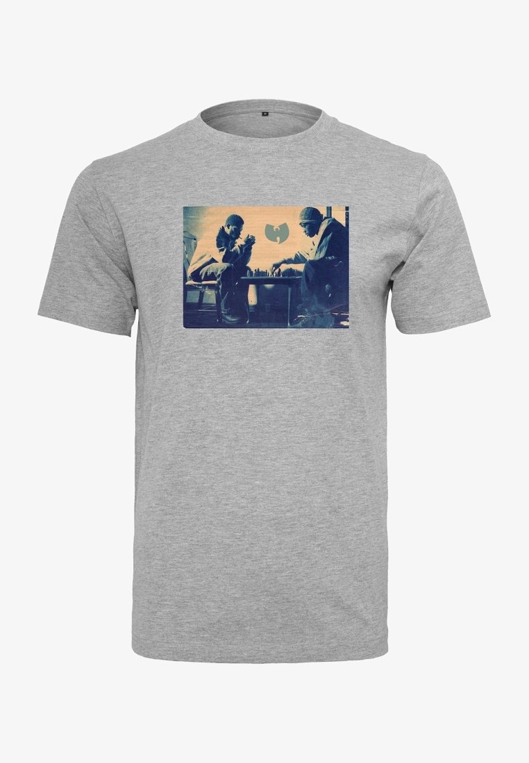 Dos hombres mirándose intensamente mientras juegan al ajedrez, con un logotipo de murciélago estilizado de fondo en una camiseta gris.