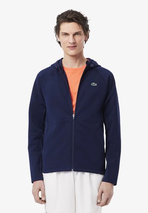Jonge man die een marineblauwe zip-up hoodie draagt met een groen krokodillenlogo, oranje t-shirt en witte broek, staand tegen een witte achtergrond.