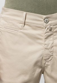 Pantalon en coton beige avec fermeture à bouton, devant plat et poche arrière simple ; présente des détails de couture et une texture minimale.