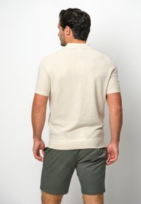 Beige Strick-Polohemd mit kurzen Ärmeln, strukturiertem Stoff und Kragen, kombiniert mit dunkelgrünen Shorts.