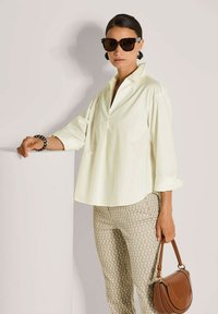 Frau mit übergroßen Sonnenbrillen, cremefarbener lockerer Bluse, gemusterten Hosen und einer braunen Ledertasche, die sich an einen weißen Sockel lehnt.