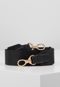 Sangle de sac en tissu noir avec un motif embossé subtil, accents en cuir noir et fermoirs pivotants en métal doré aux deux extrémités, enroulée sur une surface blanche.