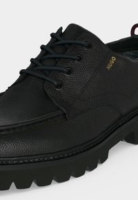 Chaussure habillée en cuir noir texturé avec semelle à gros crampons, fermeture à lacets sur l'avant et logo doré "HUGO" sur le côté.