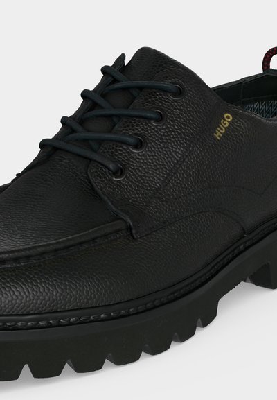 Chaussure habillée en cuir noir texturé avec semelle à gros crampons, fermeture à lacets sur l'avant et logo doré "HUGO" sur le côté.