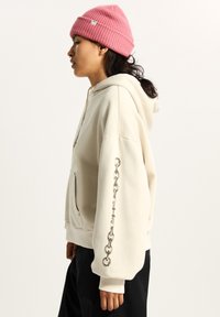 Beige hoodie met een buidelzak aan de voorkant, voorzien van een zilveren kettingafbeelding op de rechtermouw. Gepaard met een roze geribbelde beanie.