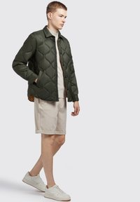 Groene gewatteerde jas met een kraag, knopen aan de voorkant, zijzakken en een gladde afwerking. Gedragen over een beige top en shorts, met lichte sneakers.