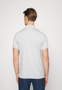 Man som vänder bort med ryggen, iklädd en ljusgrå kortärmad pikétröja och mörkblå byxor framför en helt vit bakgrund.