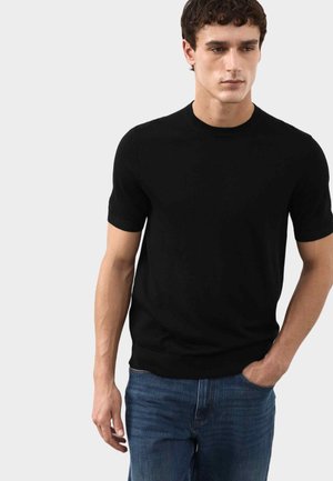 Giovane uomo con una maglietta nera aderente a maniche corte e collo rotondo, e jeans blu, con una mano in tasca, su uno sfondo chiaro e semplice.