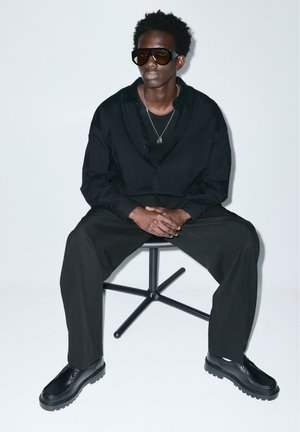 Muškarac u crnoj odjeći, s naočalama za sunce i ogrlicom, sjedi na crnoj okretnoj stolici na jednobojnoj bijeloj pozadini.