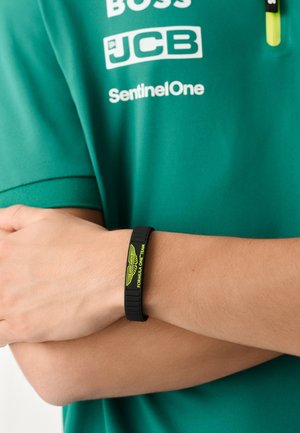 Melna gumijas aproce ar izspiestu dzeltenu uzrakstu "FORMULA ONE TEAM" un logotipu, valkāta uz rokas pret tirkīzzilas krekla fonu.