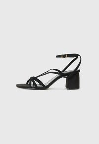 Sandale en daim noir avec un talon bloc, présentant plusieurs lanières et une boucle à la cheville. Semelle lisse et design minimaliste.
