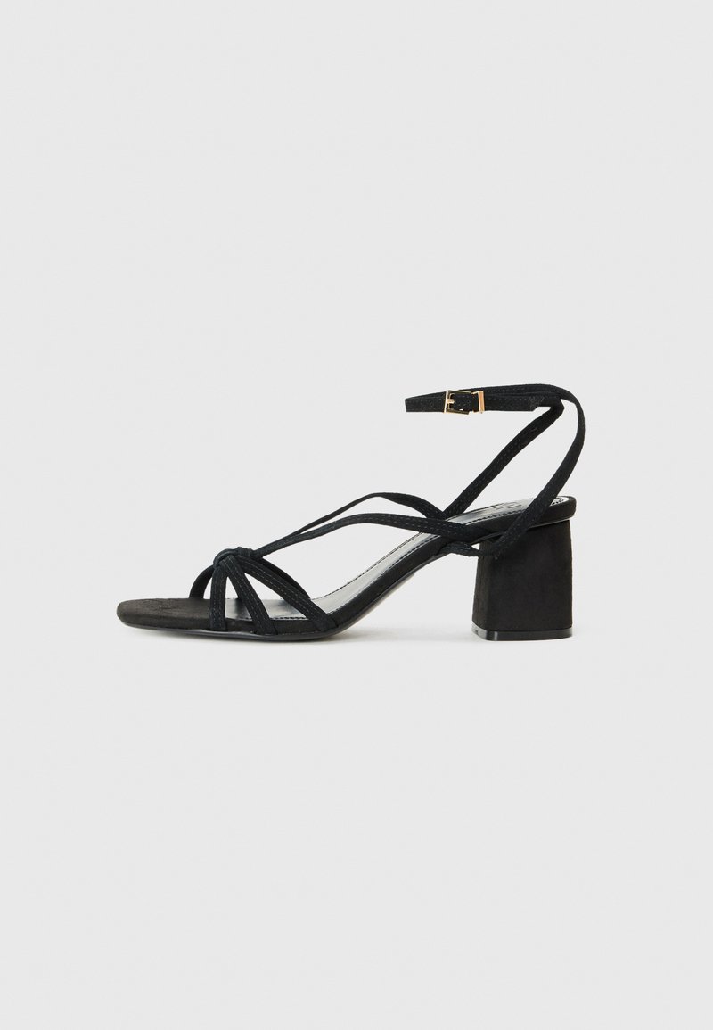 Sandale en daim noir avec un talon bloc, présentant plusieurs lanières et une boucle à la cheville. Semelle lisse et design minimaliste.