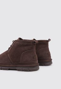 Botines de ante marrón con punta redondeada, diseño con cordones y acabado texturizado. Cuentan con suela de goma plana y detalles de marca.