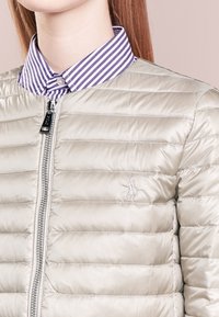 Light beige puffer jacket with quilted texture, silver zipper, and small logo; layered over a purple and white striped collared shirt.