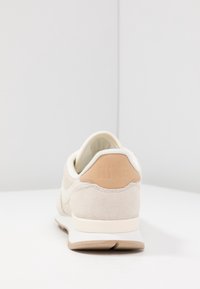 Beige mockask sneaker med en krämfärgad mesh-ovandel, med en texturerad tan-hälaccent och en vit mellansula. Nike-logotypen är präglad på hälen.