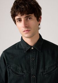 Camisa de denim verde oscuro con cuello puntiagudo, botones frontales y acentos bordados en el pecho. Presenta dos bolsillos en el pecho con solapas en ángulo.