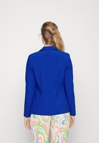 Modrý upravený blazer s jedním rozparkem vzadu, nošený přes barevné vzorované kalhoty. Hladká tkanina, strukturovaný design a klopový límec.