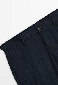 Mörkblå byxor tillverkade av ett texturerat tyg med fiskbensmönster. Har en knappstängning och bälteshällor för justerbar passform.