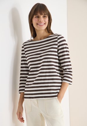 Femme souriante, portant un haut à manches longues rayé horizontalement marron et blanc et un pantalon beige clair, debout contre un mur blanc et beige.