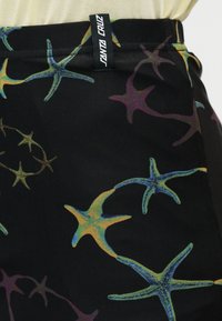 Nahaufnahme eines schwarzen Stoffes mit buntem Seesternmuster und einem schwarzen Etikett mit der Aufschrift "Santa Cruz", das am Bund angenäht ist.