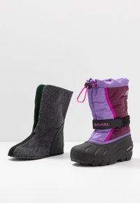 Botte d'hiver isolée violette et noire à côté de sa doublure intérieure amovible grise et verte, toutes deux posées sur une surface blanche.