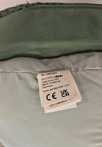 Gai zaļas auduma spilventiņš ar teksturētu malu, uz kura redzama balta etiķete ar informāciju par atbilstību un ražotāju.