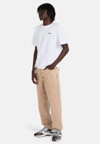 Λευκό βαμβακερό T-shirt με μικρό μαύρο λογότυπο, συνοδευόμενο από μπεζ παντελόνι φαρδιάς γραμμής και πολύχρωμα αθλητικά παπούτσια.