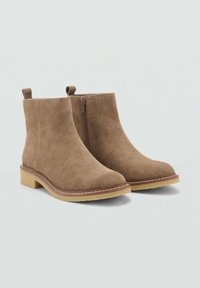 Par de botas de ante al tobillo en marrón claro con cremallera lateral, tiradores y suela de crepé beige sobre un fondo claro liso.