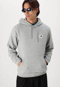 Grå hettegenser med kengurulomme, snorhette og en rund logo-patch. Båret over en blå skjorte, tilpasset mørke shorts.