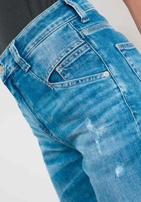 Gros plan sur une personne portant un jean en denim déchiré bleu clair avec un haut noir rentré à l'intérieur.