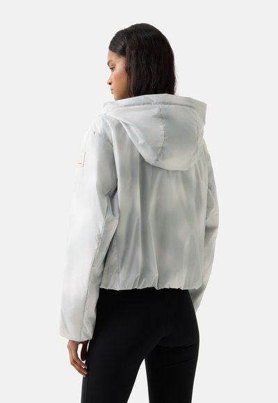 Vrouw met lang donker haar, gekleed in een lichtgrijze gewatteerde jas met capuchon en zwarte legging, zijwaarts kijkend tegen een witte achtergrond.