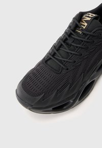 Black athletic sneaker with textured knit upper, unique sculpted sole with hollow sections, and gold branding on the tongue.