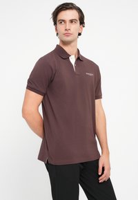 Hnědá polokošile s bílým límečkem, krátkými rukávy a postranními rozparky. Na hrudi vyšívané logo "HACKETT". Hladká struktura látky.