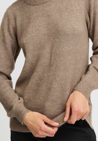 Jasnobrązowy sweter z dzianiny z wysokim kołnierzem, ściągaczami przy mankietach i miękką teksturą. Dół jest nieco dłuższy z tyłu, o luźnym kroju.