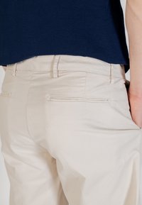 Pantalons en coton beige avec poches avant et taille plate, présentant des détails de coutures subtils et une coupe décontractée. Haut bleu marine partiellement visible.