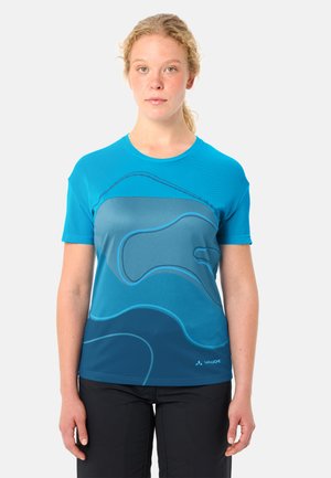 Junge Frau mit lockigem blondem Haar, die ein blau gemustertes kurzärmeliges Sportshirt und schwarze Hosen trägt, vor einem einfarbigen Hintergrund steht.