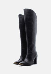 Ψηλές μαύρες δερμάτινες μπότες με μυτερές χρυσές μεταλλικές μύτες και ψηλά τετράγωνα τακούνια, απεικονίζονται όρθιες σε λευκό φόντο.