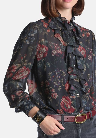 Femme portant un chemisier noir transparent à motif floral avec un devant à volants, ceinture bordeaux, jean foncé et bracelet manchette en cuir noir avec détail doré.