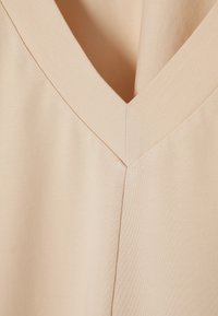 Beige V-nyakú ruhadarab, sima felülettel, tiszta varrással és diszkrét fényességgel. Az anyag puha esést biztosít, hangsúlyozva a minimalista dizájnt.