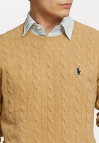 Pull en maille torsadée de couleur jaune moutarde, fabriqué en matériau doux, avec un col rond et un logo brodé sur la poitrine.