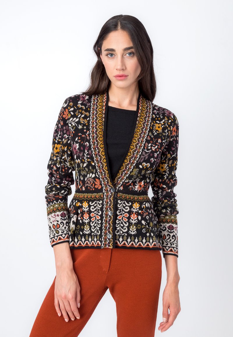 Cardigan nero con ricami floreali colorati, tessuto testurizzato, scollo a V, chiusura con bottoni e maniche lunghe. Abbinato a pantaloni arancioni.