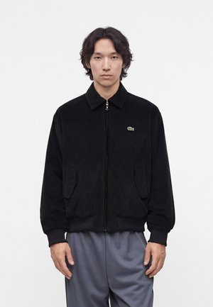 Giovane uomo con capelli di media lunghezza che indossa una giacca nera con cerniera e logo e pantaloni grigi, in piedi davanti a uno sfondo bianco semplice.
