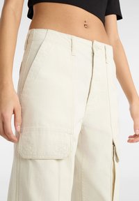 Wijdvallende cargobroek in lichtbeige stof met frontzakken en gestikte details. Kenmerkt zich door een hoge taille en een subtiele glans.