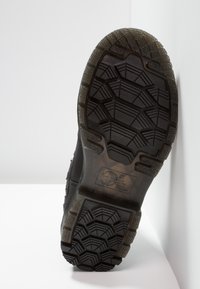 Svart skosåle med teksturert mønster, med dype spor for grep. Gummimateriale med logodetaljer og sklisikre egenskaper.